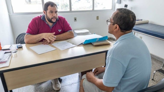 Profissional de saúde realiza atendimento a paciente durante consulta em unidade de saúde de Jundiaí, com prontuários sobre a mesa e ambiente clínico ao fundo.