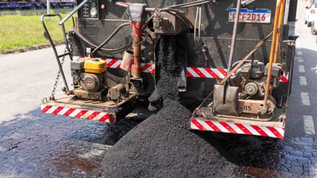 Caminhão da manutenção viária aplica massa asfáltica em serviço de tapa-buracos em rua urbana.