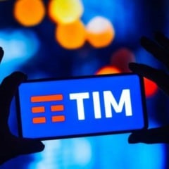 Pessoa segura um celular com o logotipo da TIM na tela, em ambiente com ilumina&ccedil;&atilde;o desfocada em tons de azul e amarelo.