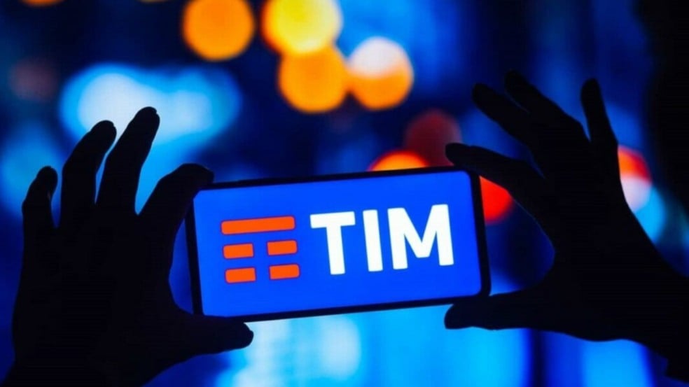 Pessoa segura um celular com o logotipo da TIM na tela, em ambiente com iluminação desfocada em tons de azul e amarelo. Pessoa segura um celular com o logotipo da TIM na tela, em ambiente com iluminação desfocada em tons de azul e amarelo.
