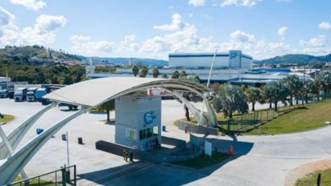 Fachada da unidade industrial da Ypê com portaria, galpões e caminhões, cercada por áreas verdes e morros ao fundo.