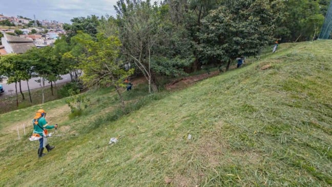 Trabalhadores realizam roçagem em área verde pública de Jundiaí, em terreno inclinado próximo a bairros residenciais.