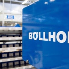 Equipamento industrial azul com o logotipo da B&ouml;llhoff em destaque, em ambiente fabril com prateleiras de pe&ccedil;as ao fundo.