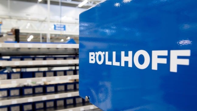 Equipamento industrial azul com o logotipo da Böllhoff em destaque, em ambiente fabril com prateleiras de peças ao fundo.