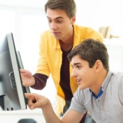 Dois jovens trabalhando juntos em frente a um computador em ambiente corporativo, com um deles apontando para a tela durante uma orienta&ccedil;&atilde;o.