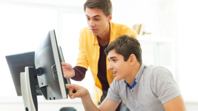 Dois jovens trabalhando juntos em frente a um computador em ambiente corporativo, com um deles apontando para a tela durante uma orientação.