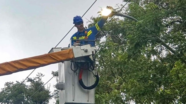 Trabalhador faz substituição de luminária por LED em poste de iluminação pública usando caminhão com cesto aéreo.