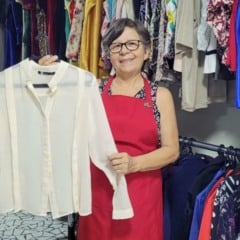 Volunt&aacute;ria do Bazar do Hospital S&atilde;o Vicente segura uma camisa branca em meio a araras com roupas femininas, em espa&ccedil;o organizado do bazar solid&aacute;rio.