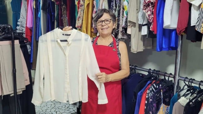 Voluntária do Bazar do Hospital São Vicente segura uma camisa branca em meio a araras com roupas femininas, em espaço organizado do bazar solidário.