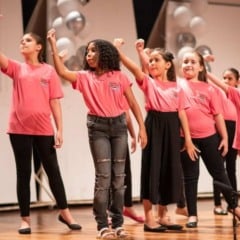 Crian&ccedil;as vestidas com camisetas rosas se apresentam em coral infantil no palco, realizando movimentos coreografados durante a apresenta&ccedil;&atilde;o.