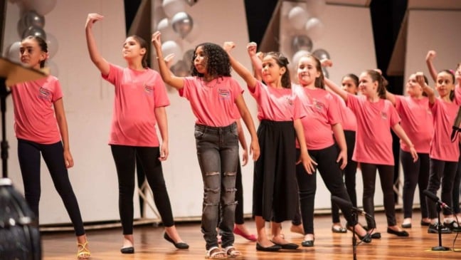 Crianças vestidas com camisetas rosas se apresentam em coral infantil no palco, realizando movimentos coreografados durante a apresentação.