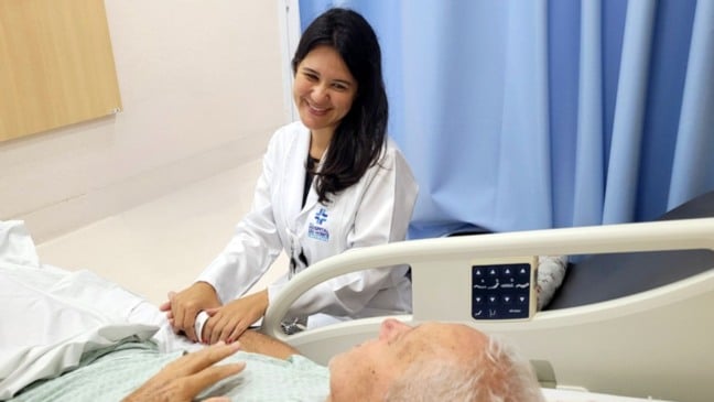 Médica do Hospital São Vicente segura a mão de paciente idoso em leito hospitalar, demonstrando cuidado humanizado e acolhimento durante atendimento de saúde.