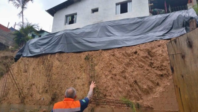 Agente da Defesa Civil avalia encosta com lona de proteção sob casa em área de risco de deslizamento após chuvas.