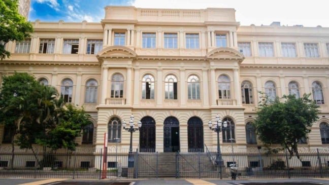 Fachada de prédio histórico da Secretaria da Educação do Estado de São Paulo, com arquitetura clássica, janelas em arco, escadaria central e vegetação ao redor.