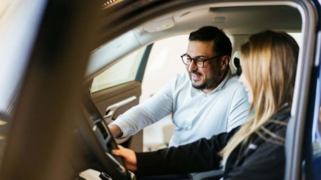 Vendedor de concessionária orienta cliente dentro de carro durante atendimento de vendas.