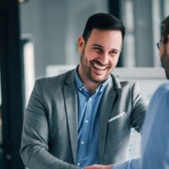 Dois homens em escrit&oacute;rio apertam as m&atilde;os sorrindo, em clima profissional, sugerindo acordo ou contrata&ccedil;&atilde;o.