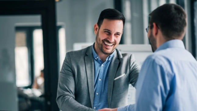Dois homens em escritório apertam as mãos sorrindo, em clima profissional, sugerindo acordo ou contratação.