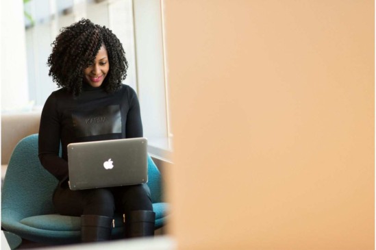 Mulher sentada em uma poltrona usando um notebook, em ambiente interno moderno e iluminado.