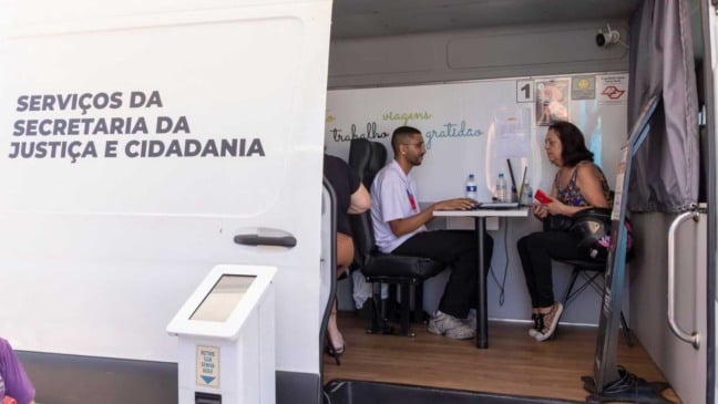 Atendimento da Secretaria da Justiça e Cidadania dentro de unidade móvel, com moradora sendo orientada por atendente durante ação de serviços públicos em bairro.