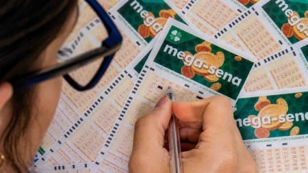 Pessoa preenche volante da Mega-Sena com caneta sobre uma mesa cheia de bilhetes da loteria. Pessoa preenche volante da Mega-Sena com caneta sobre uma mesa cheia de bilhetes da loteria.